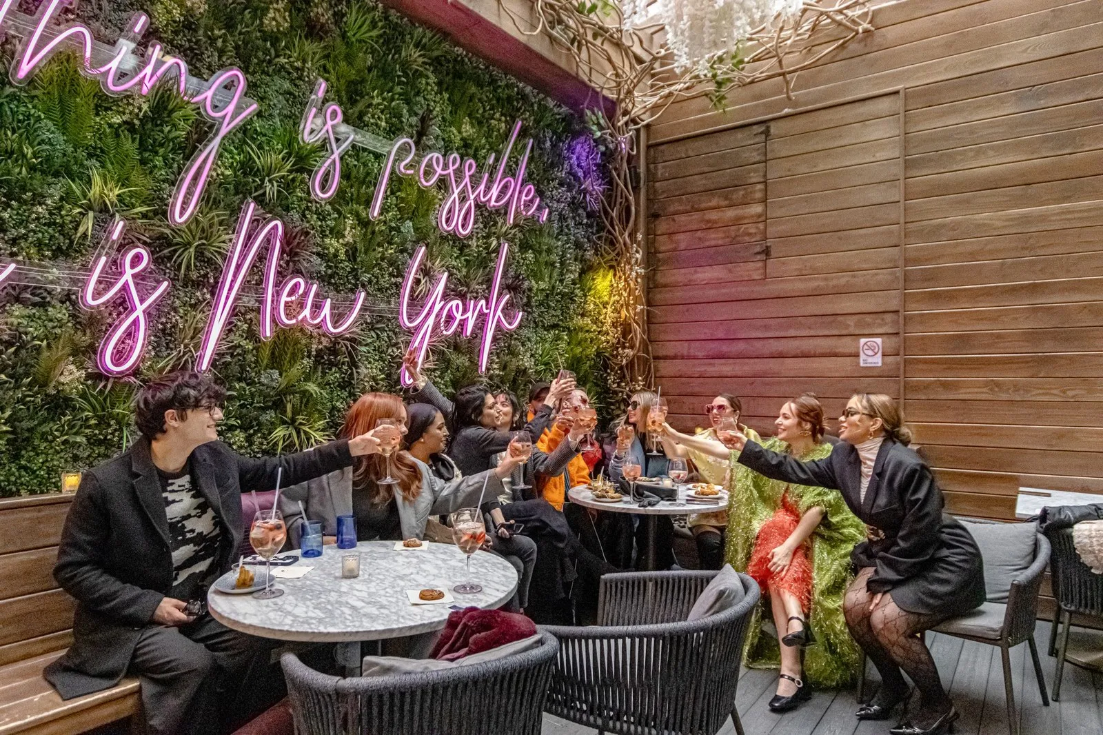 Guests raising their glasses around a table on a hotel terrace, under a pink neon sign reading “Anything is possible, this is New York”