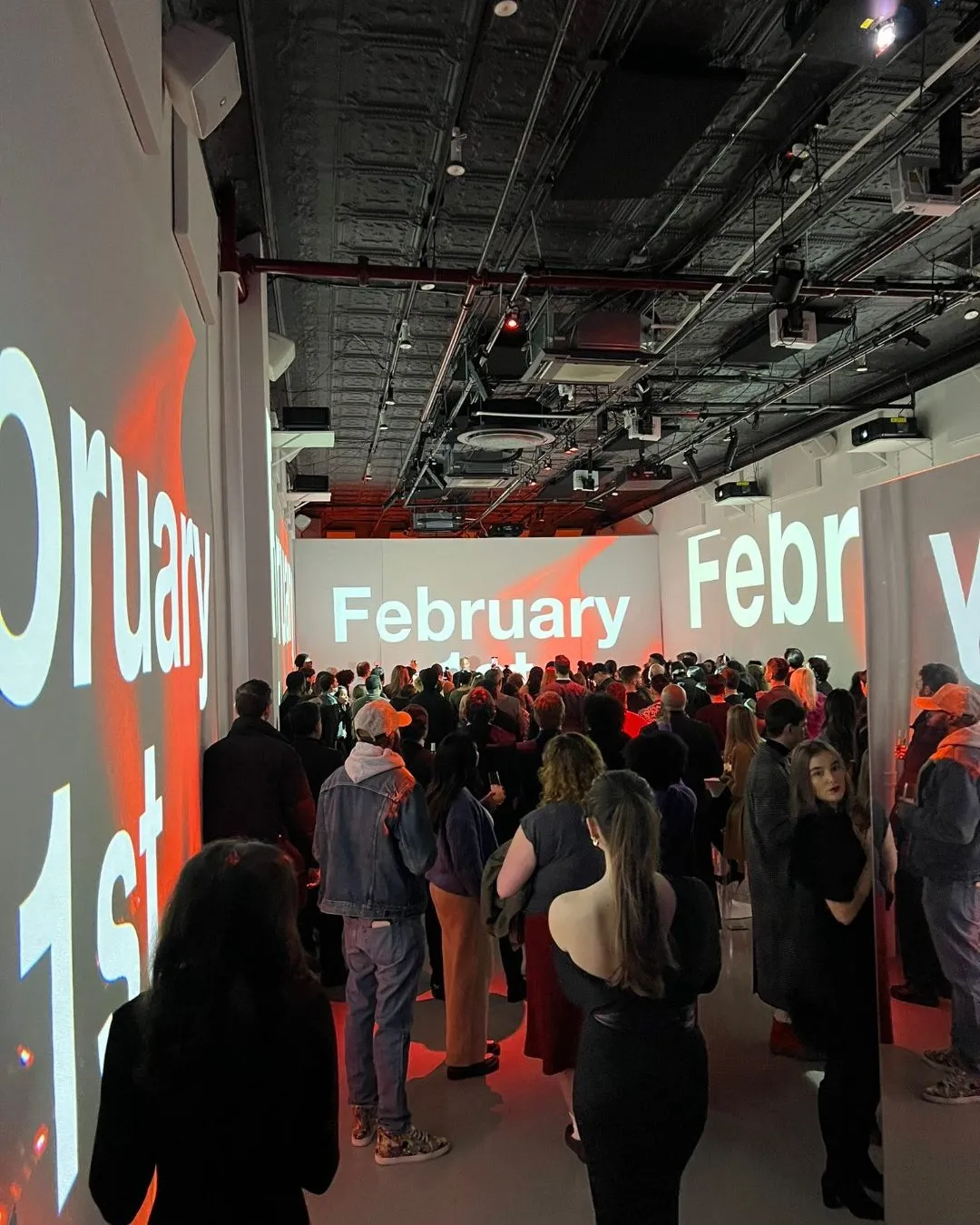 A crowd gathered in a dark industrial venue under projected text reading “February 1st”