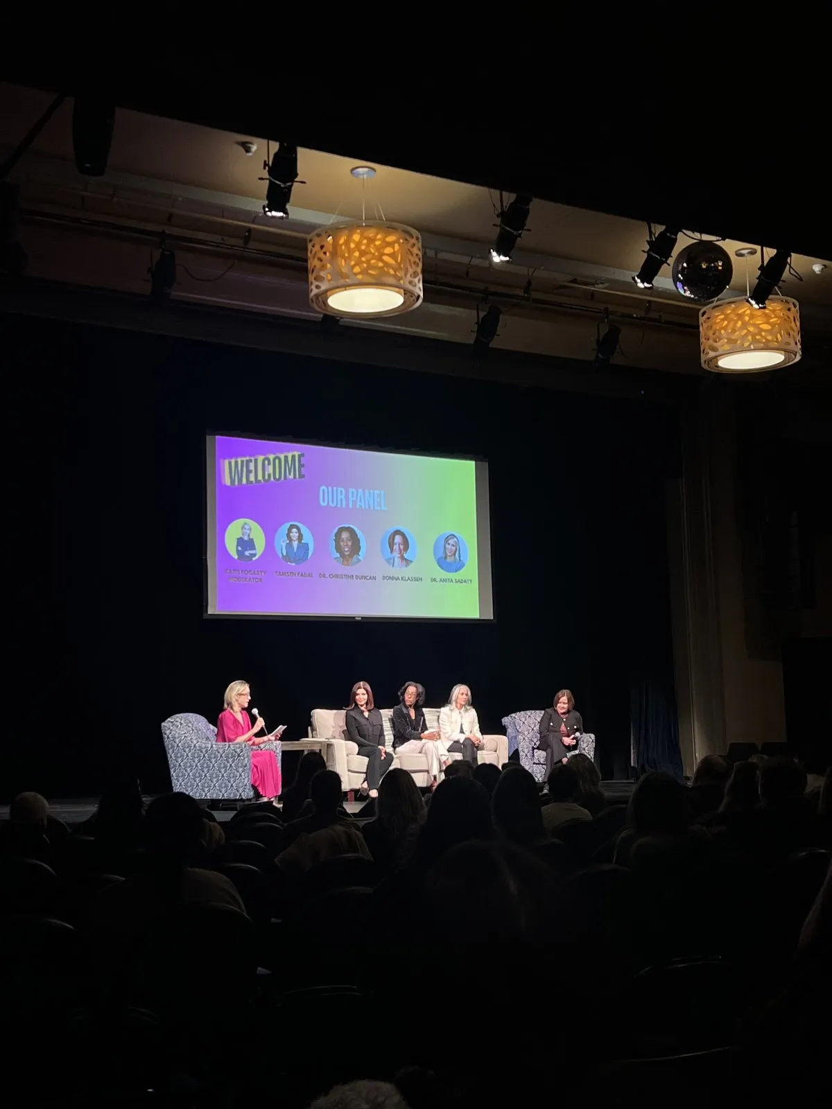 Five speakers seated on a dark stage below a screen reading “Welcome” and “Our panel”, with the audience in silhouette
