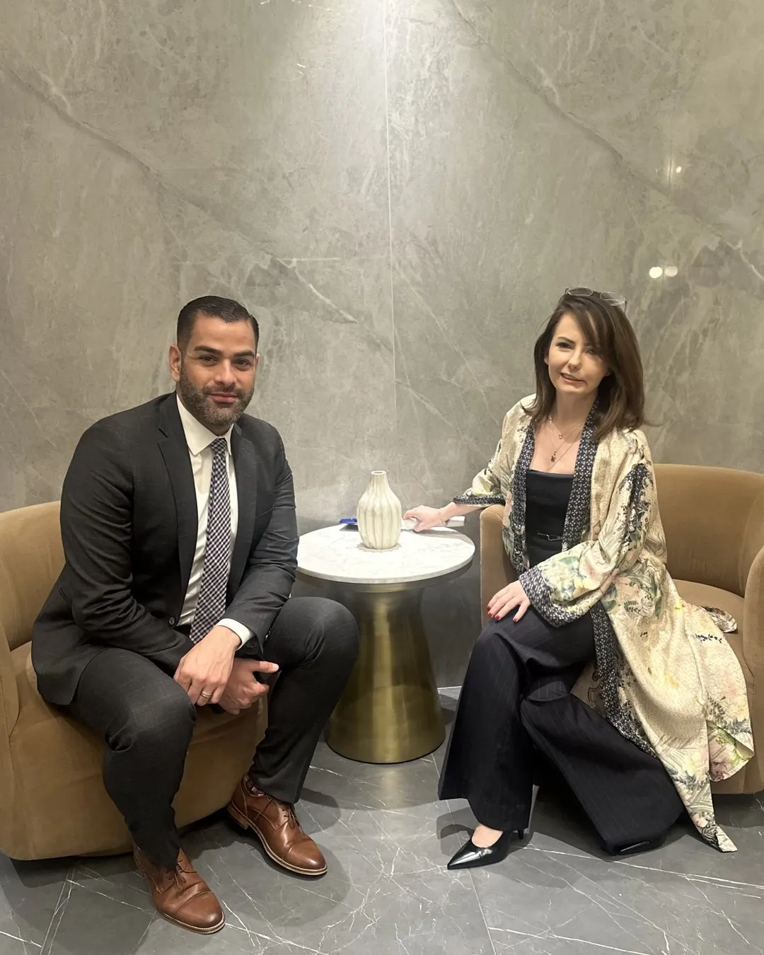 A man in a suit and a woman in a patterned robe seated facing each other in a hotel lobby