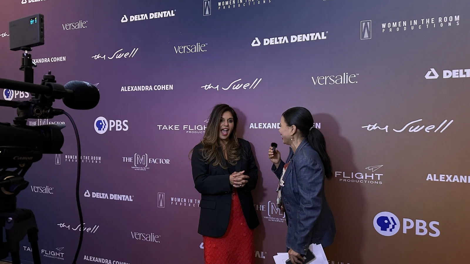A woman being interviewed in front of a camera at a press wall covered in sponsor logos