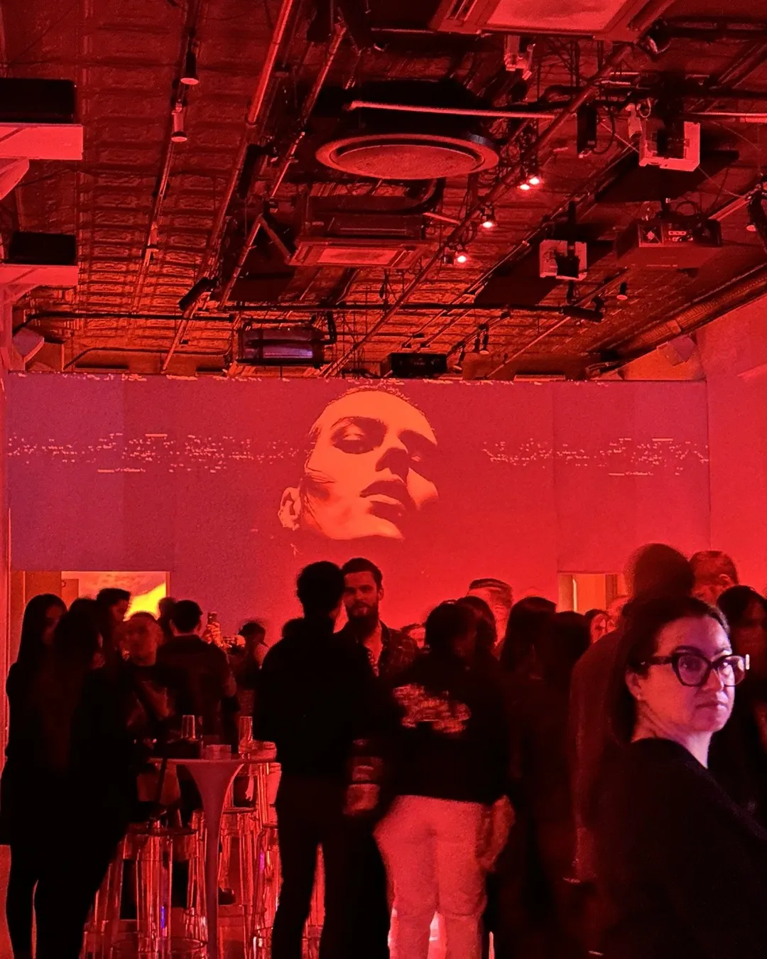 Guests in a venue lit in deep red, with a large face projected on the wall